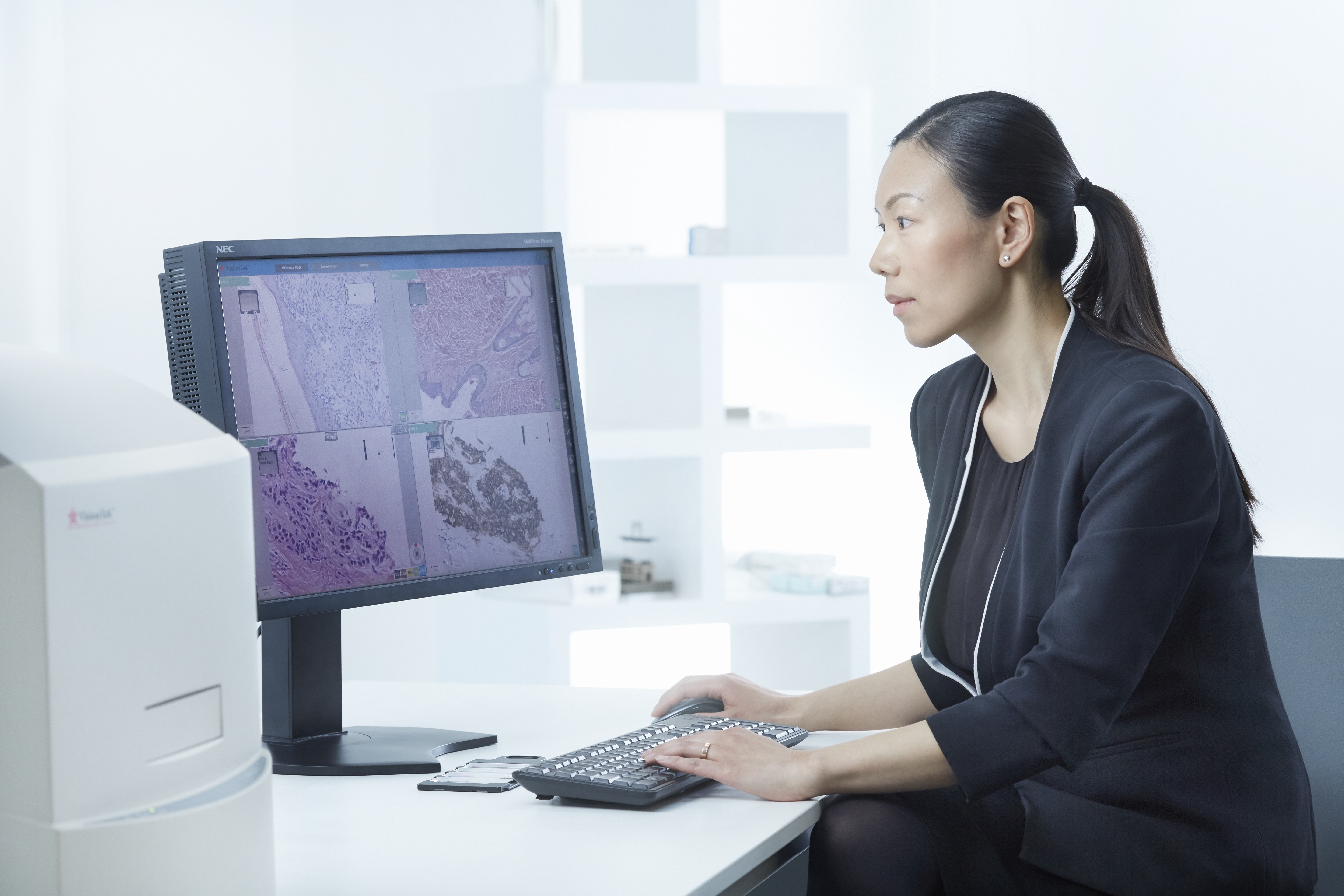 A woman in business attire examines tissue sample images on a computer monitor in a modern office setting.