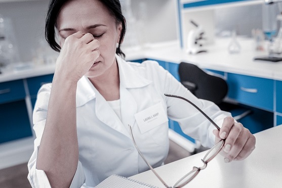 A person in a lab coat sits at a desk, eyes closed and pinching the bridge of their nose while holding glasses, appearing tired or stressed in a laboratory setting.