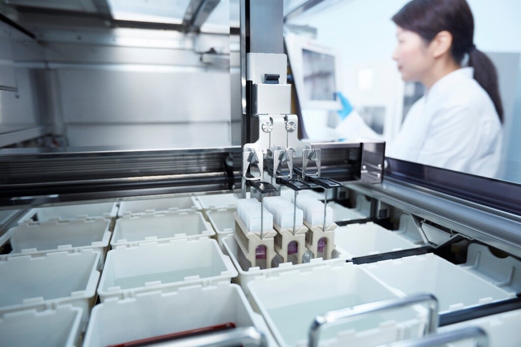 A laboratory automation machine handles test samples, with a researcher working at a computer in the background.