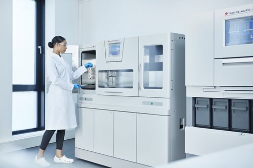A scientist in a lab coat and gloves places a sample tray into a large automated laboratory machine in a bright laboratory room.