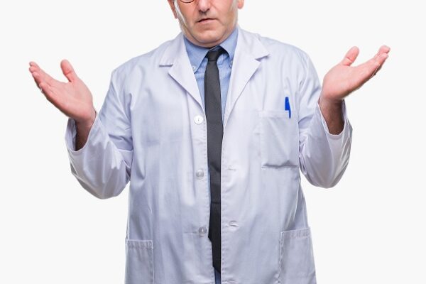 A man in a white lab coat and tie shrugs with his hands raised and an uncertain expression on his face, standing against a plain white background.