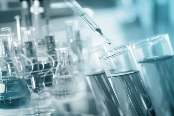 Close-up of a pipette dispensing liquid into a test tube, with several flasks and beakers containing fluids in the background, representing a laboratory setting.