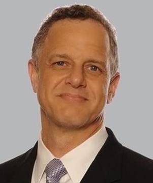 A middle-aged man in a suit and tie is smiling slightly in front of a plain gray background.
