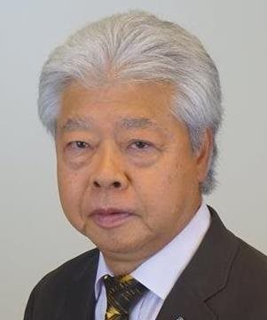 An older man with white hair and a suit poses against a plain, light-colored background.