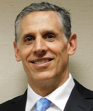 A middle-aged man with short gray hair, wearing a dark suit, white shirt, and blue patterned tie, smiling at the camera against a plain beige background.