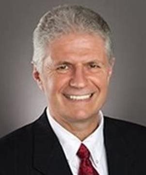 A middle-aged man with short gray hair, wearing a dark suit, white shirt, and red tie, smiling at the camera against a plain background.