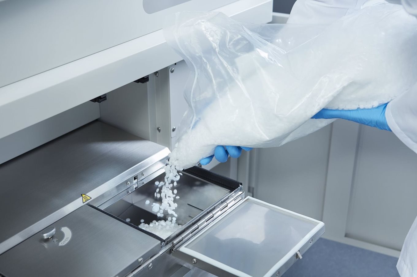 A person wearing blue gloves pours white plastic pellets from a bag into a compartment of an industrial machine.