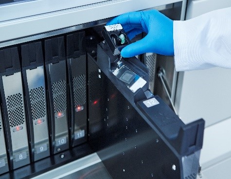 A gloved hand places a component into a laboratory instrument with numbered slots and indicator lights.