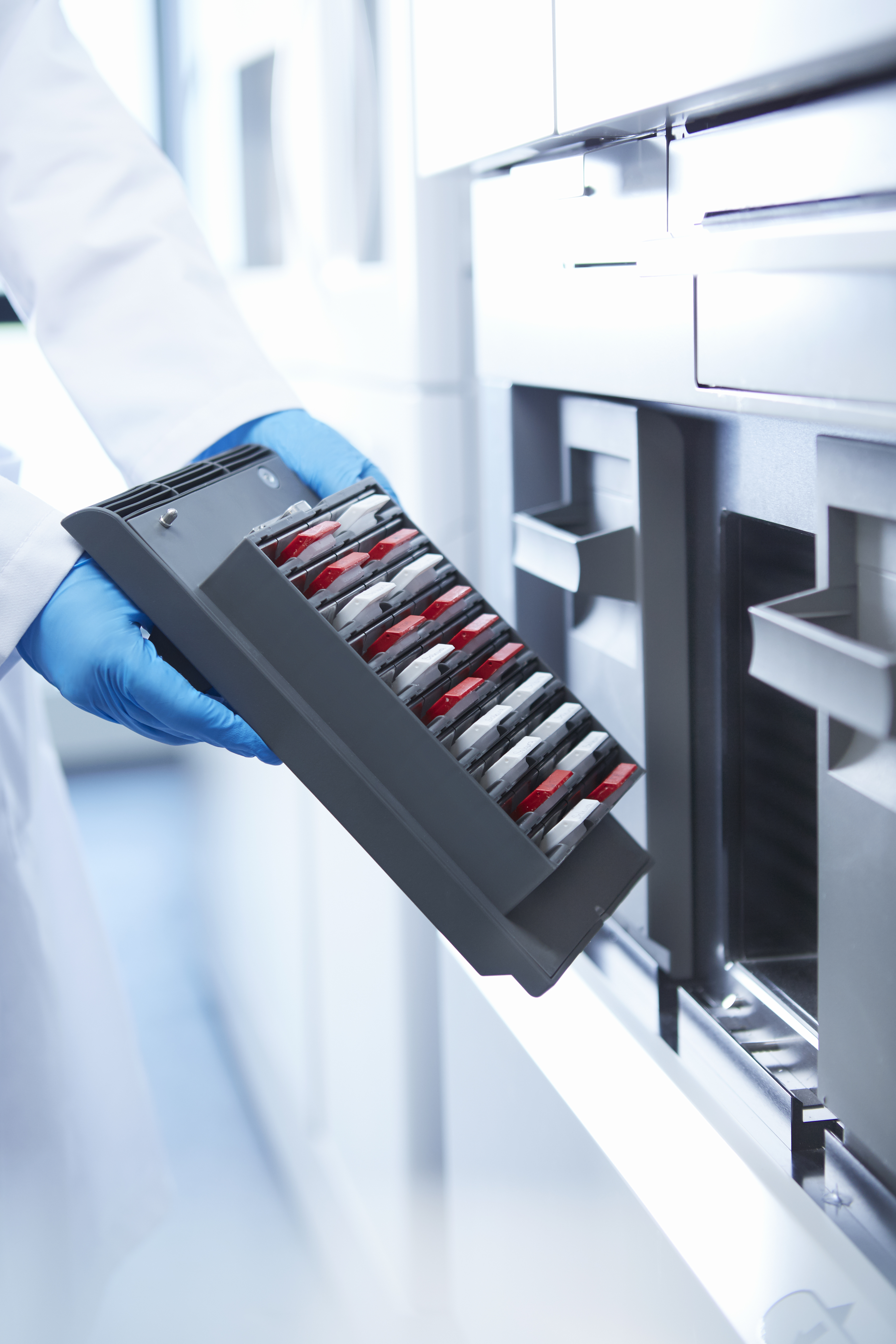 A person wearing blue gloves inserts a tray of vials or sample containers into a laboratory analysis machine.