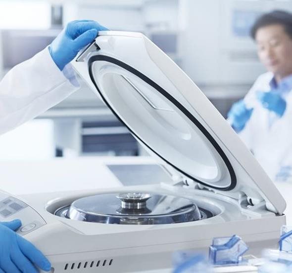 A gloved hand opens the lid of a laboratory centrifuge, with another person in a lab coat visible in the blurred background.