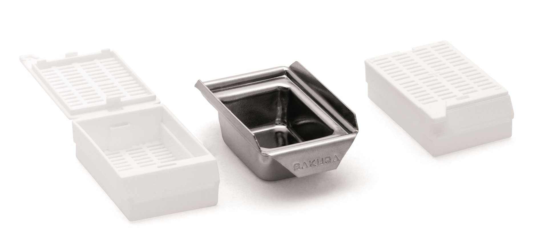 A metal tissue capsule is centered between two semi-transparent plastic tissue cassettes, one open on the left and one closed on the right.