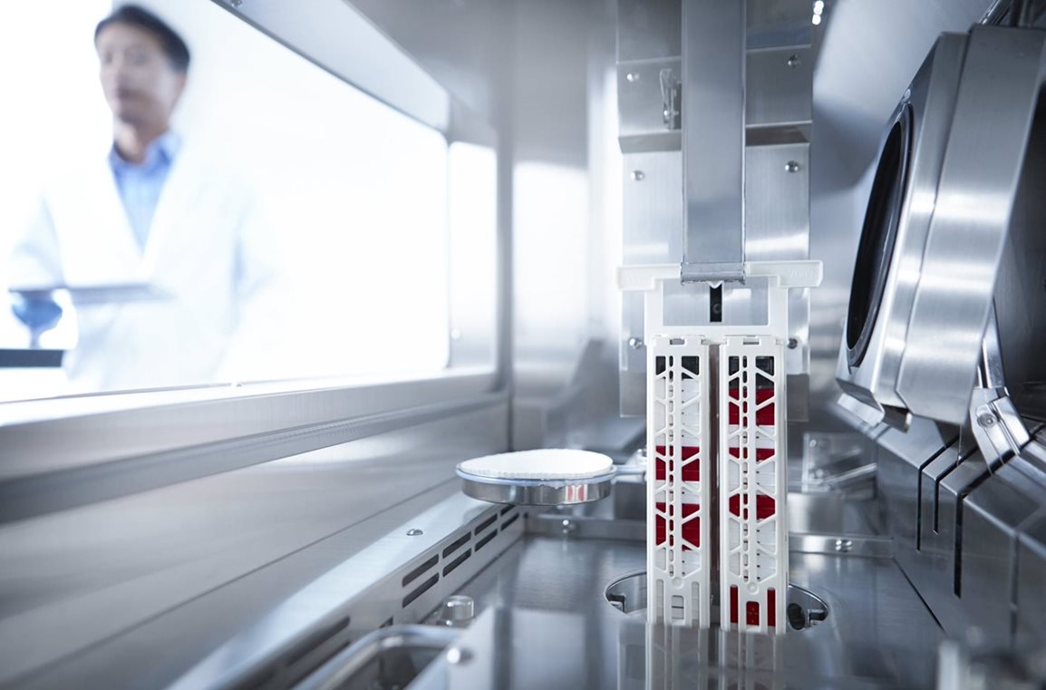 A laboratory setting with a scientist in the background and scientific equipment, including a mechanical arm holding a white rack with red samples, in the foreground.