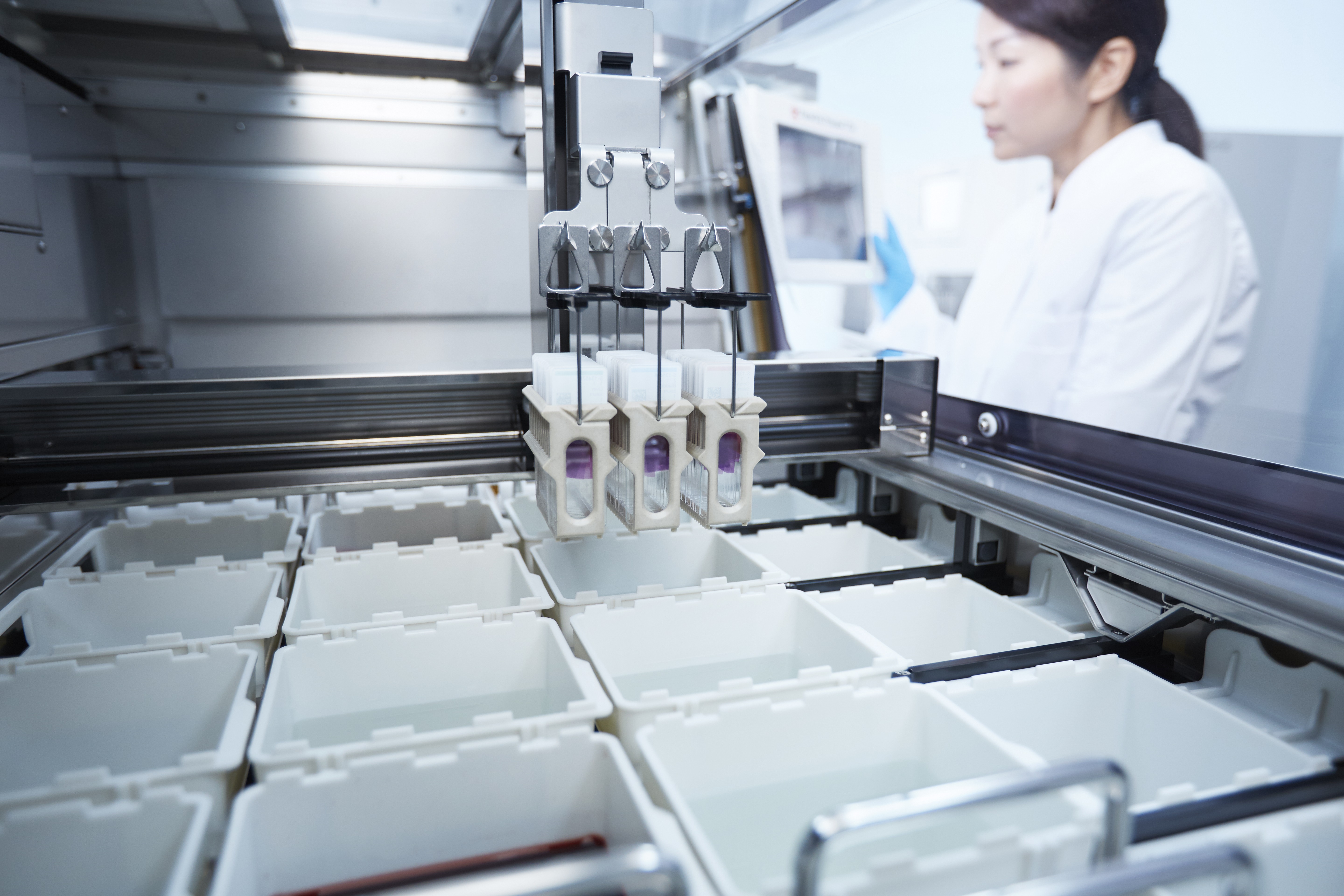 A robotic machine places containers into slots, while a person in a lab coat stands in the background monitoring the process.