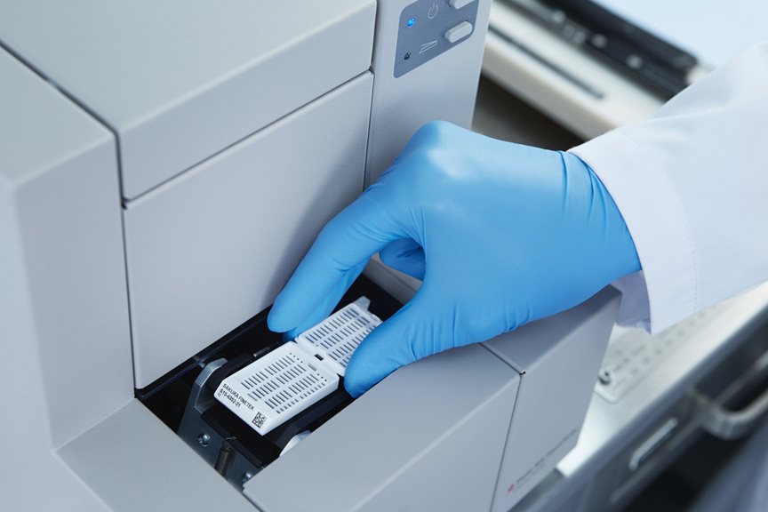 A gloved hand places a labeled sample cassette into a laboratory machine for processing or analysis.