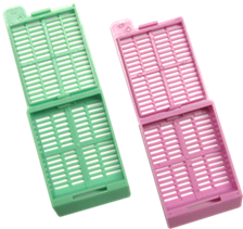 Green and pink plastic tissue cassettes with hinged lids open, showing the slotted surfaces used for holding biopsy samples in laboratories.