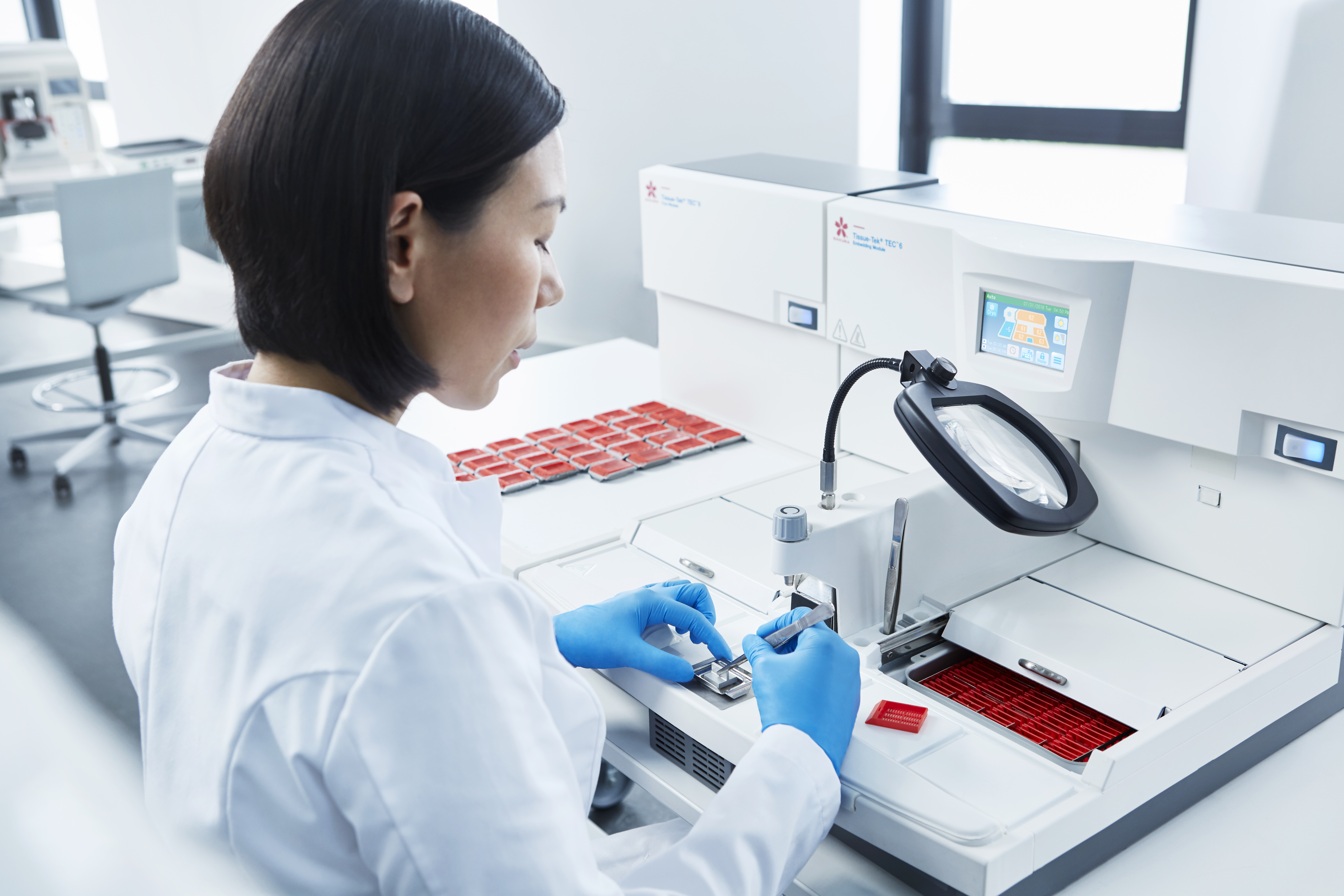 A laboratory technician in a white coat and blue gloves operates scientific equipment, handling samples on a tray in a modern lab setting.