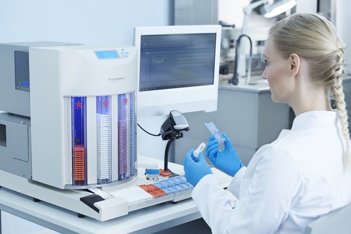 A lab technician in a white coat operates laboratory equipment and handles sample trays while working at a computer station in a modern laboratory.