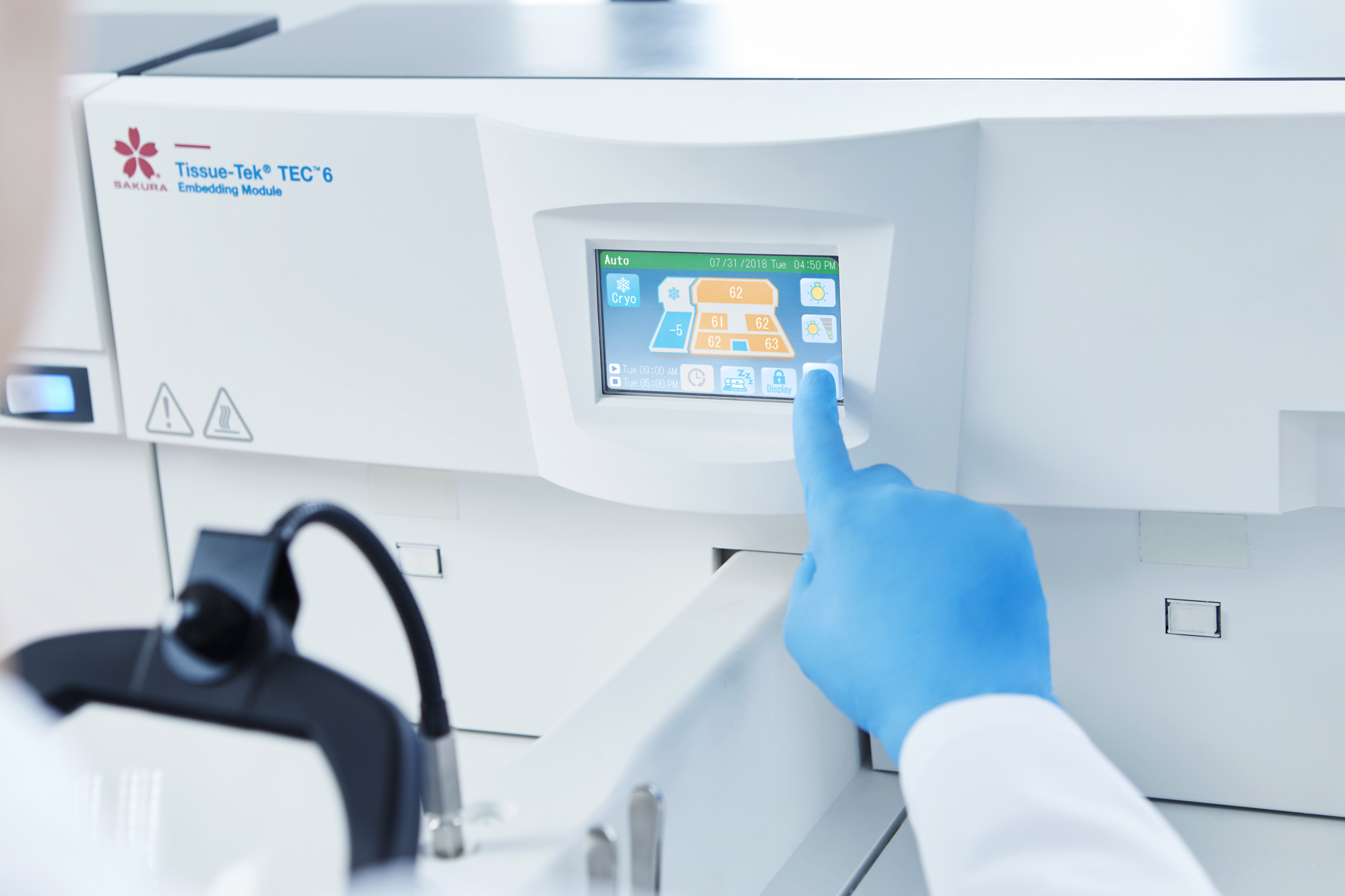 A person wearing a blue glove operates a touchscreen control panel on a laboratory device labeled