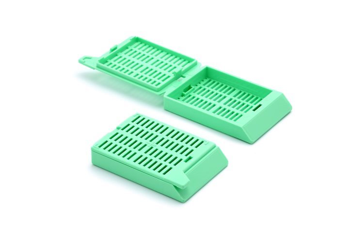 Green plastic tissue cassettes, one open and one closed, used for holding biological samples during histology processing, on a white background.