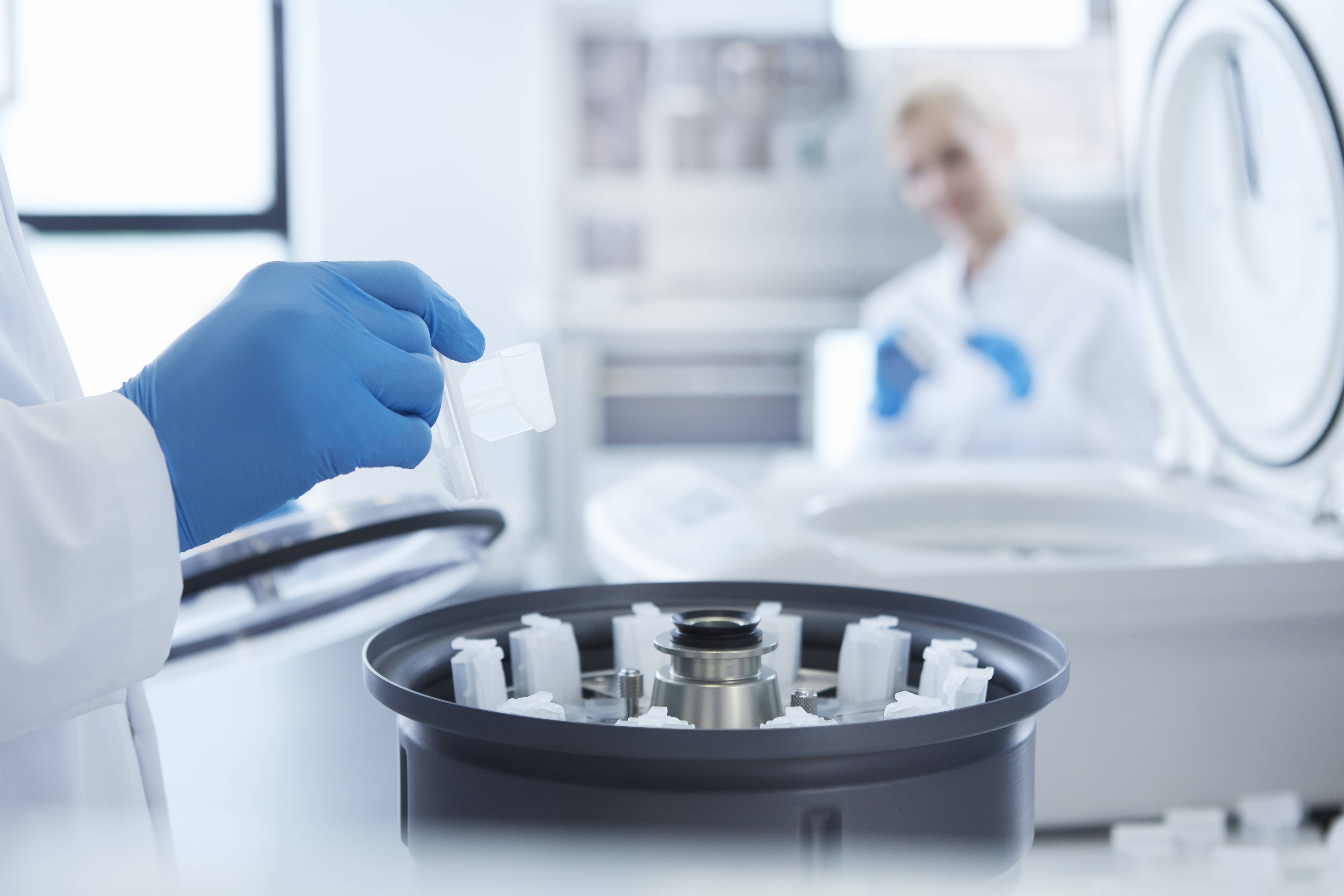 A person wearing blue gloves places a sample into a laboratory centrifuge, with another person in the background.