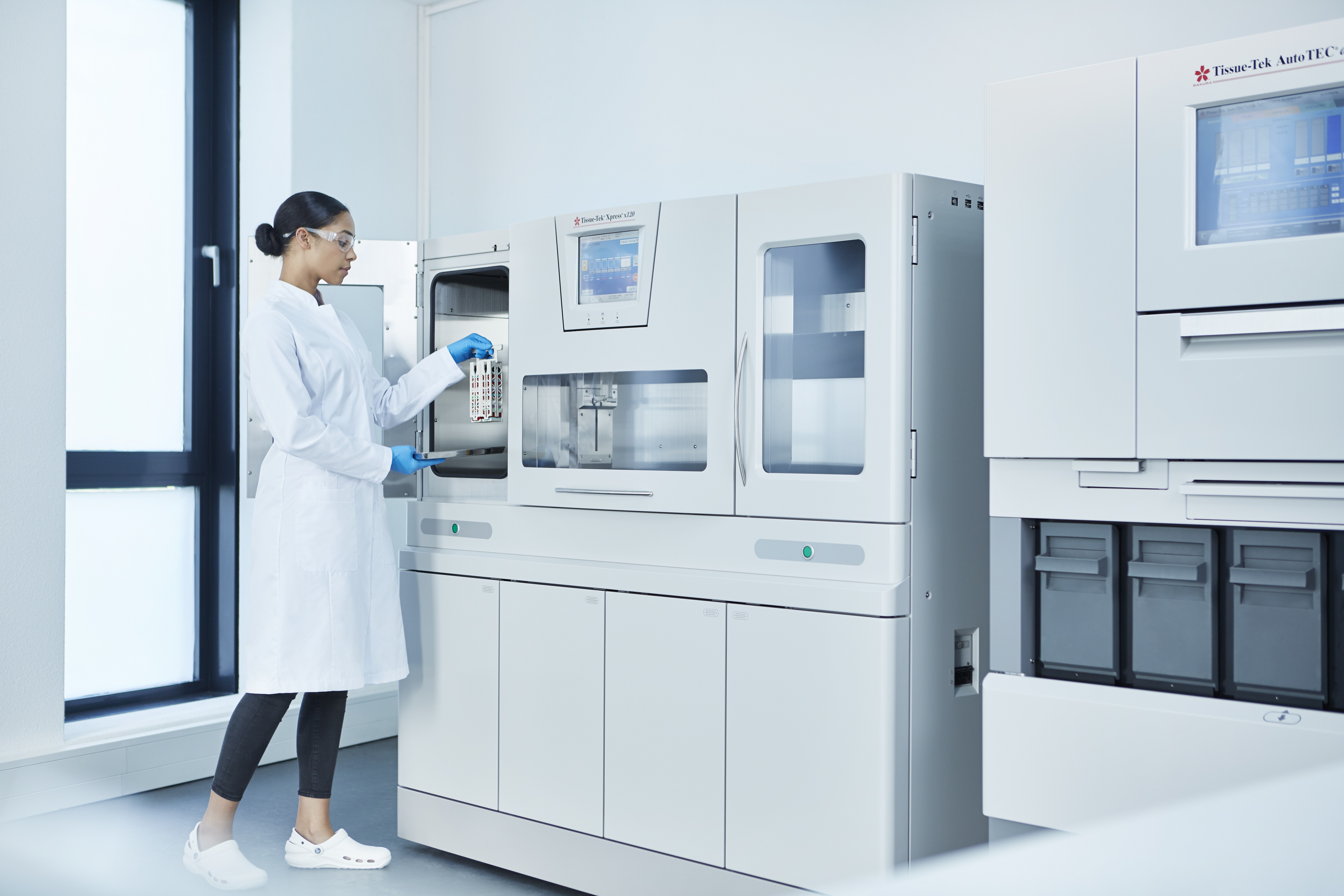A person in a lab coat and gloves loads a tray of samples into an automated laboratory analyzer in a bright, modern lab.