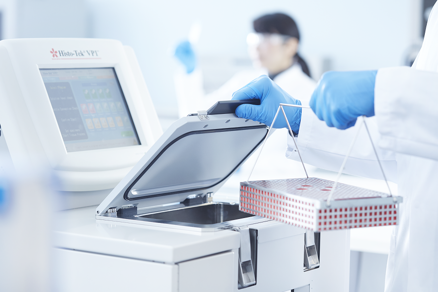 A lab technician in blue gloves places a tray of sample containers into a laboratory machine labeled Histo-Tek VP1. Another person is blurred in the background.