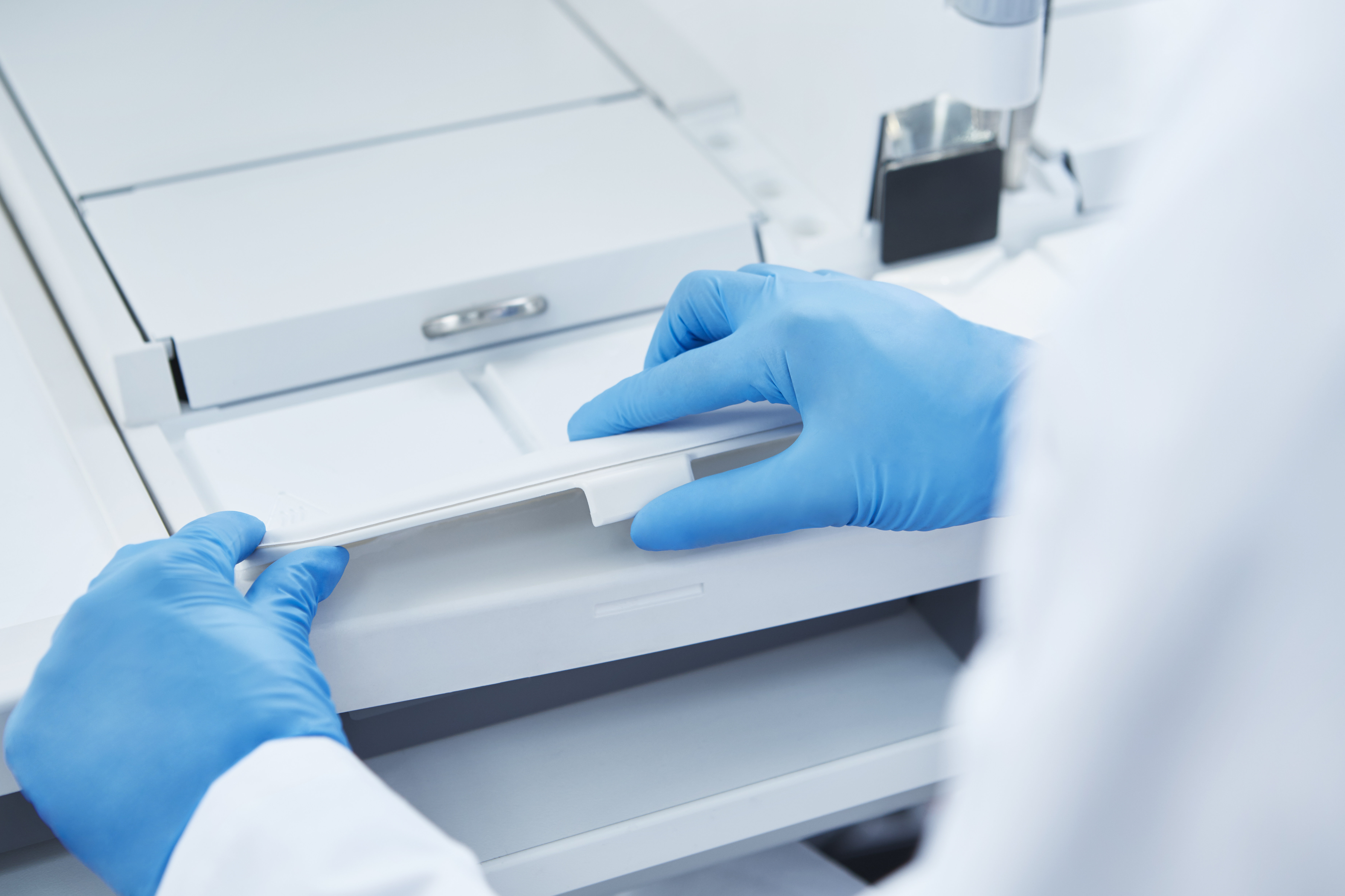 A person wearing blue gloves operates laboratory equipment, opening or closing a white compartment.