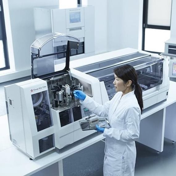 A lab technician in protective clothing operates a large automated analyzer machine in a modern laboratory setting.