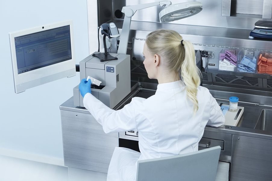 A lab technician in a white coat operates laboratory equipment at a workstation, scanning a sample with a barcode reader next to a computer monitor.