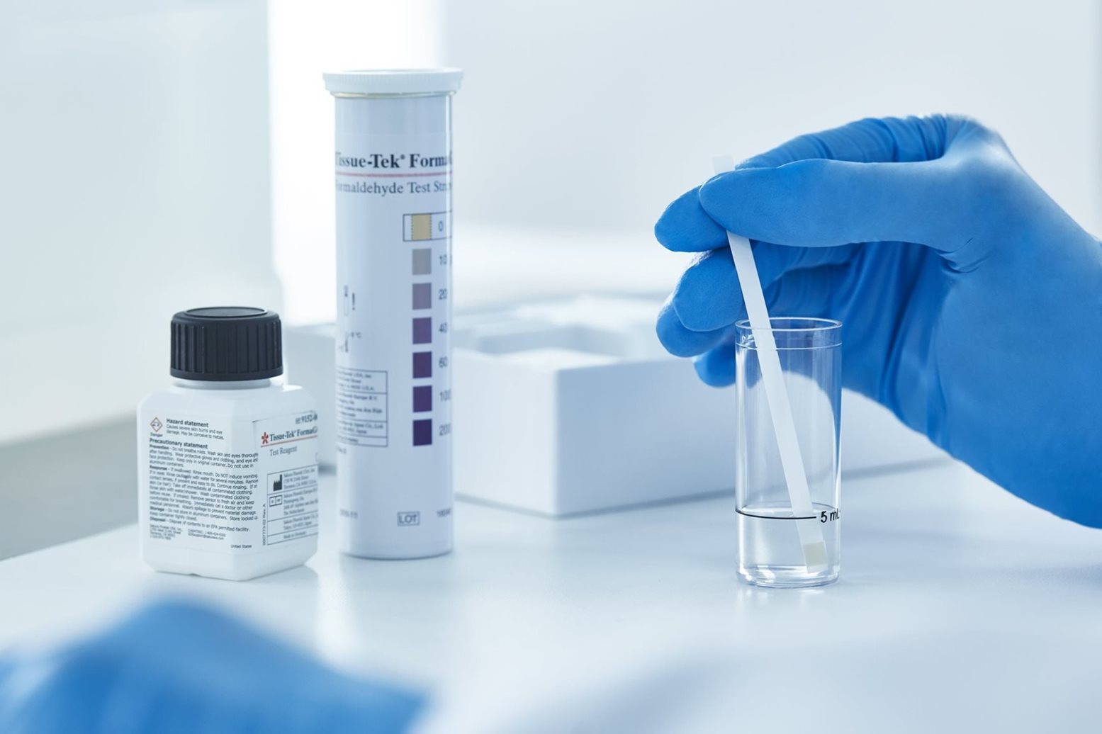 A gloved hand dips a test strip into a clear liquid in a small glass vial, with chemical bottles and a color chart in the background on a laboratory table.