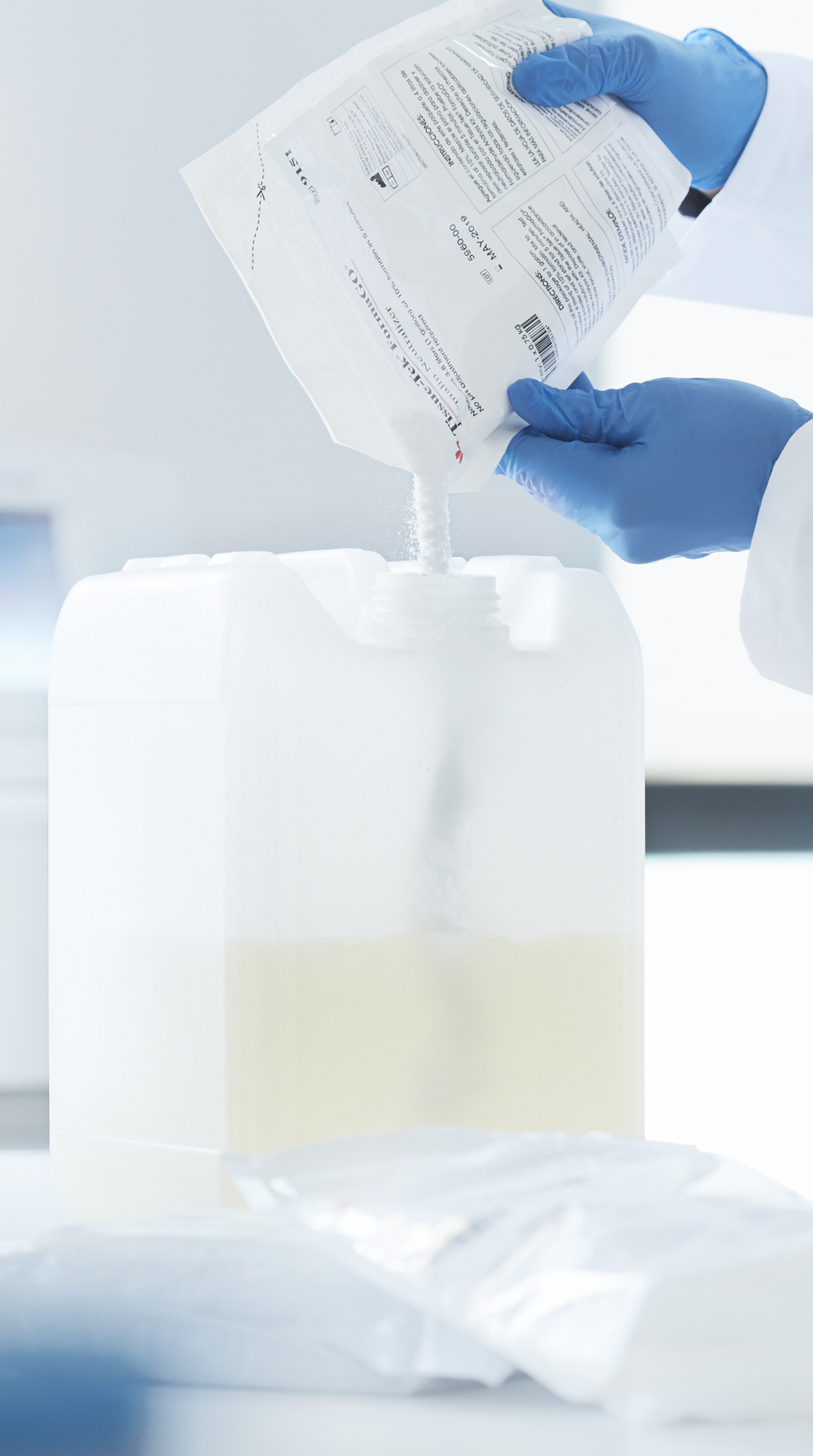 A person wearing blue gloves pours powder from a packet into a large white plastic container filled with liquid in a laboratory setting.