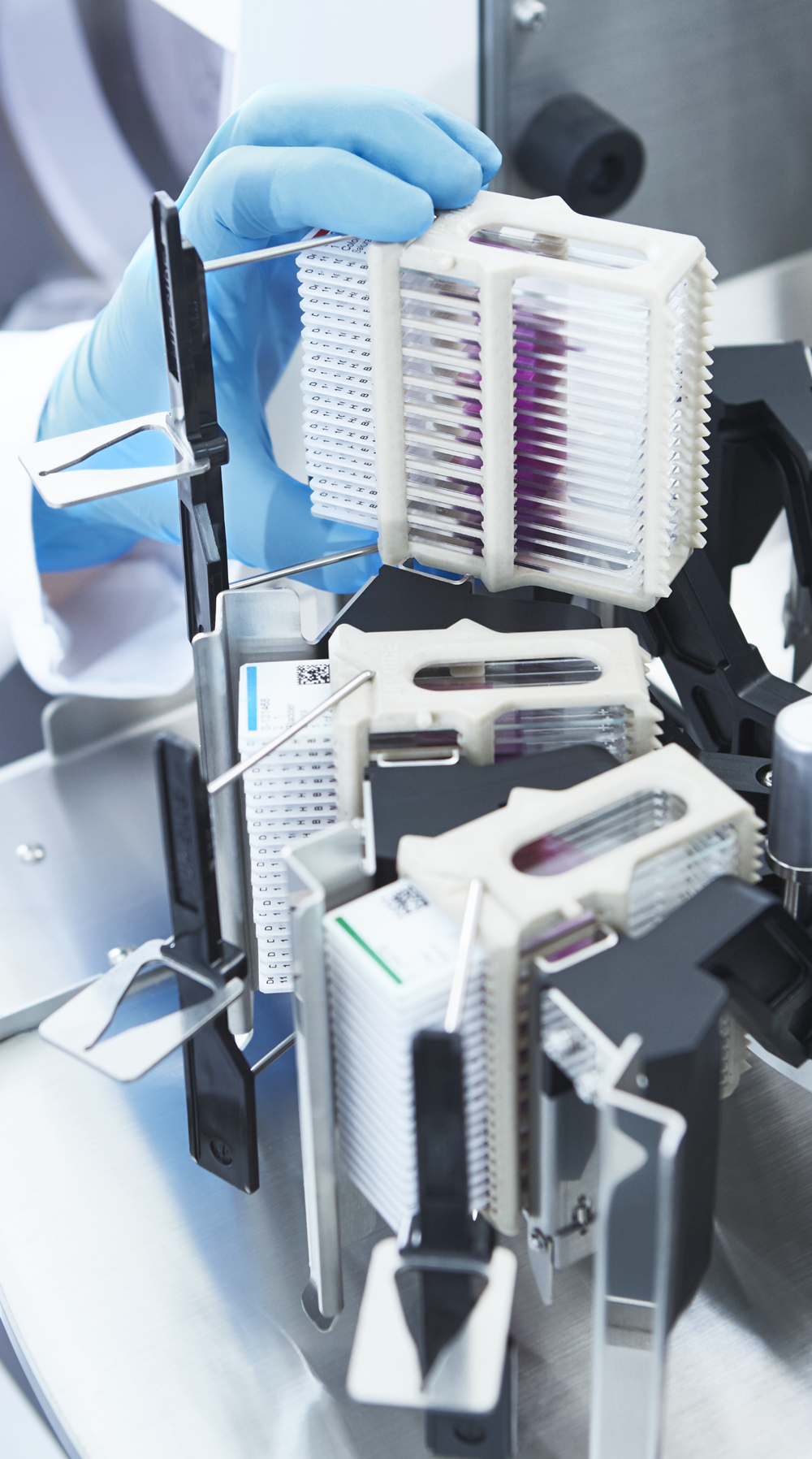 A gloved hand handles a rack of test tubes filled with liquid, placed in a laboratory machine for automated analysis.