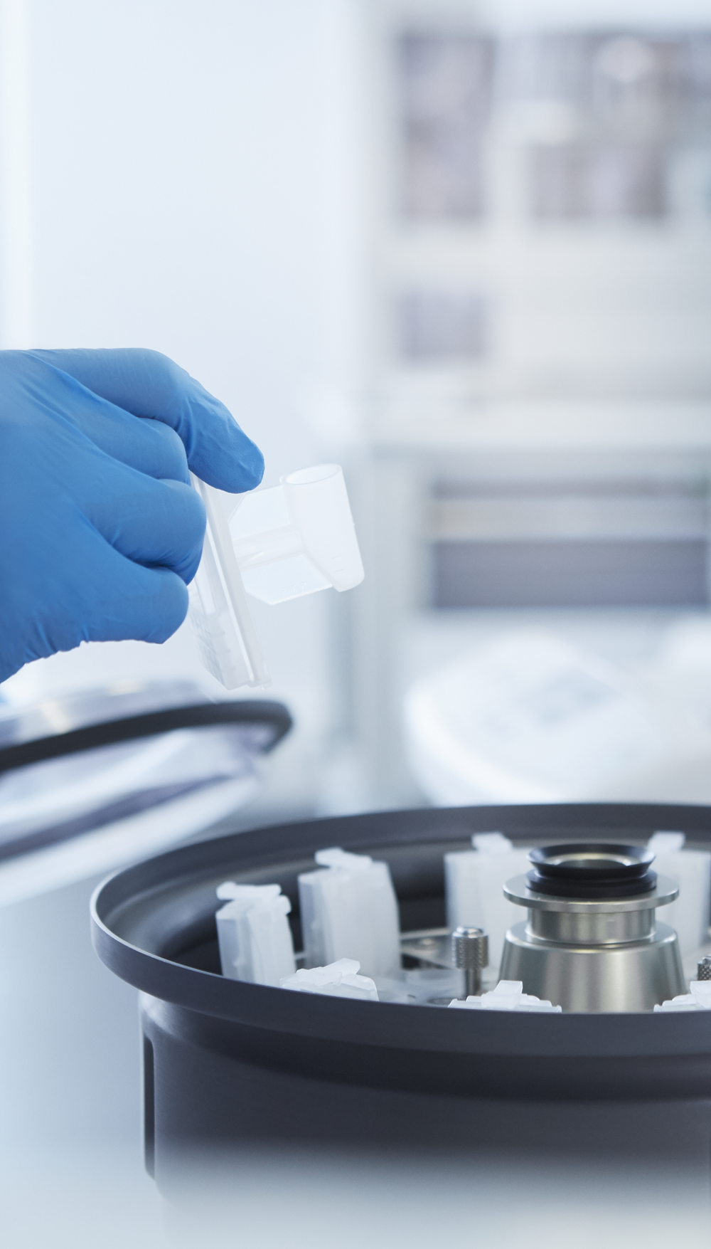A gloved hand places a sample container into a centrifuge machine in a laboratory setting.