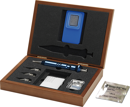 A wooden box containing a blue handheld device, metal tools, small blades, a pack of blotting papers, and a foil pouch labeled “Tissue-Tek” arranged in foam compartments.