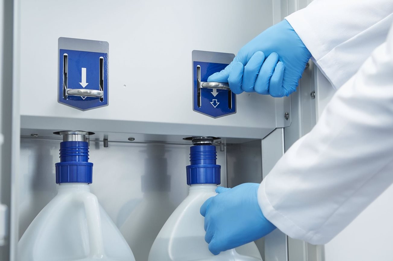 A person wearing blue gloves operates a dispensing mechanism above two large white plastic containers with blue caps in a laboratory setting.