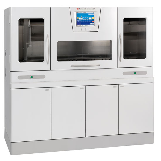 A large laboratory instrument with a central touchscreen, glass viewing panels, and storage cabinets below, used for automated sample processing.