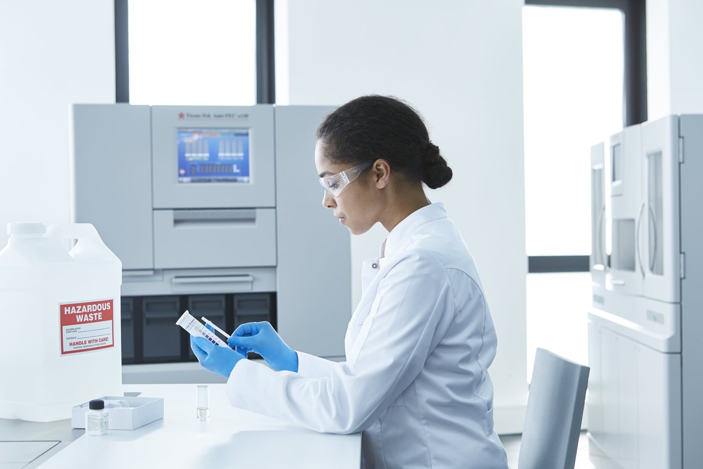 A scientist in a lab coat, gloves, and goggles examines a test tube while seated at a laboratory bench with scientific equipment and hazardous waste container nearby.