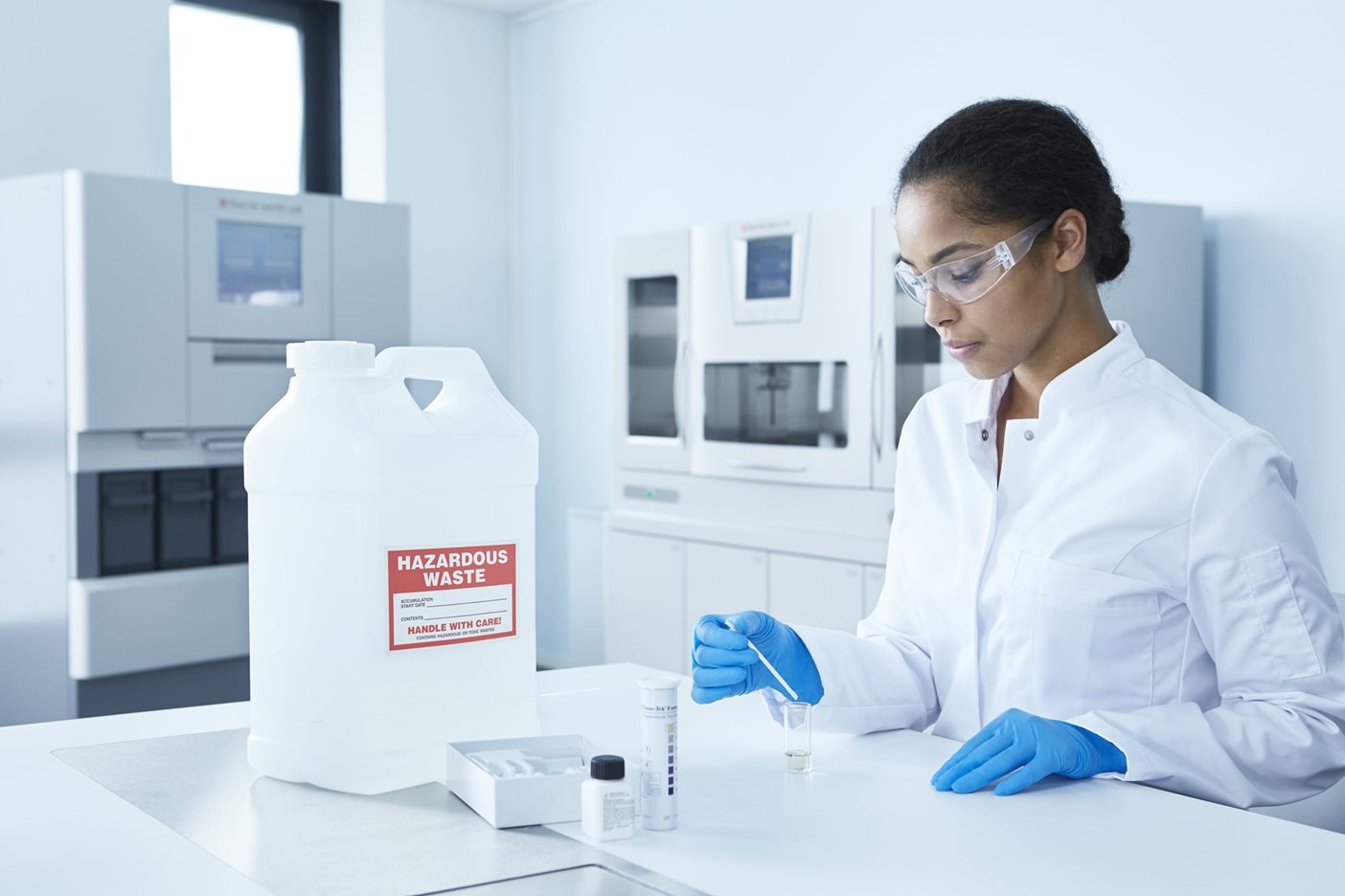 A scientist in a lab coat and gloves handles samples next to a large white container labeled