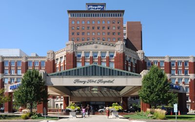 Exterior view of Henry Ford Hospital, featuring a multi-story brick building with an entrance canopy and people walking near the front.