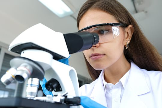 A woman wearing safety glasses and a lab coat observes a sample through a microscope in a laboratory setting.