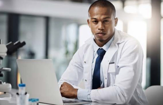 A scientist in a white lab coat sits at a desk with a laptop and laboratory equipment, looking at the camera.