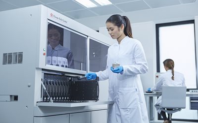 A woman in a lab coat and gloves operates scientific equipment, while another person works at a desk in the background of a laboratory.