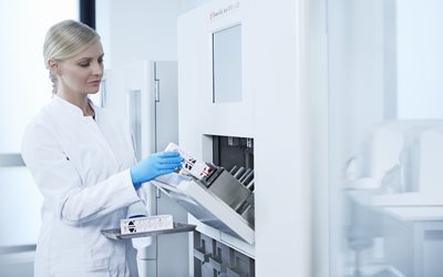 A lab technician in a white coat and blue gloves loads medical samples into a laboratory machine.