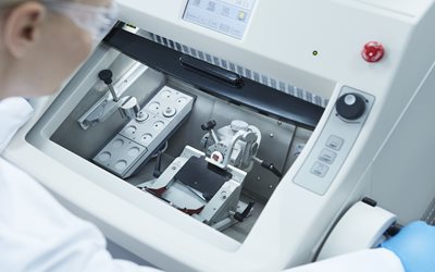 A lab technician operates a cryostat machine, preparing thin tissue samples for examination. The device’s controls and compartments are visible.