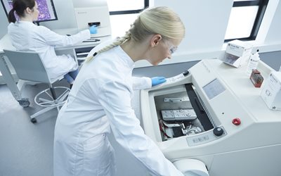 Two scientists in a laboratory; one operates a cryostat machine while the other works at a computer workstation in the background.