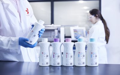 Two people in lab coats work in a laboratory; one holds a white bottle while four similar bottles with colored caps are arranged on a counter in the foreground.