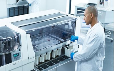 A scientist in a lab coat and gloves operates a large automated laboratory machine, placing samples into designated slots in a clinical or research laboratory setting.