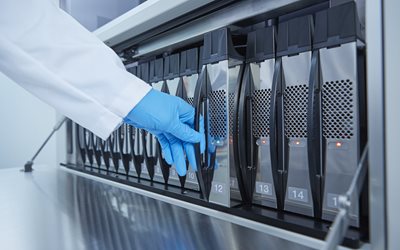 A gloved hand inserts or removes a numbered cartridge from a row of slots in a laboratory machine.