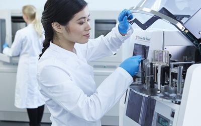A scientist wearing a lab coat and blue gloves operates a laboratory machine, while another person works in the background.