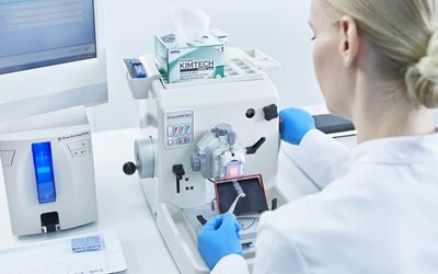 A lab technician wearing blue gloves uses a microtome to slice a sample for analysis in a laboratory setting.